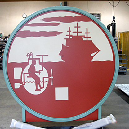Round metal sign featuring cut-out silhouettes of a person sitting inside a pipe and a ship on the horizon with clouds above