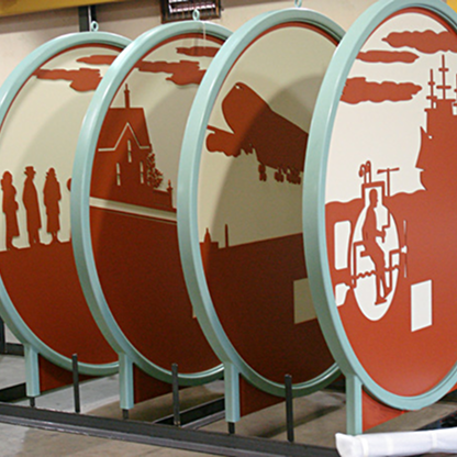 Series of large round metal signs with artistic cut-out designs depicting silhouettes of people, a plane, a house, and industrial imagery.