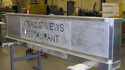 Custom metal sign with engraved text 'GRACIE MEWS RESTAURANT' displayed in a workshop.